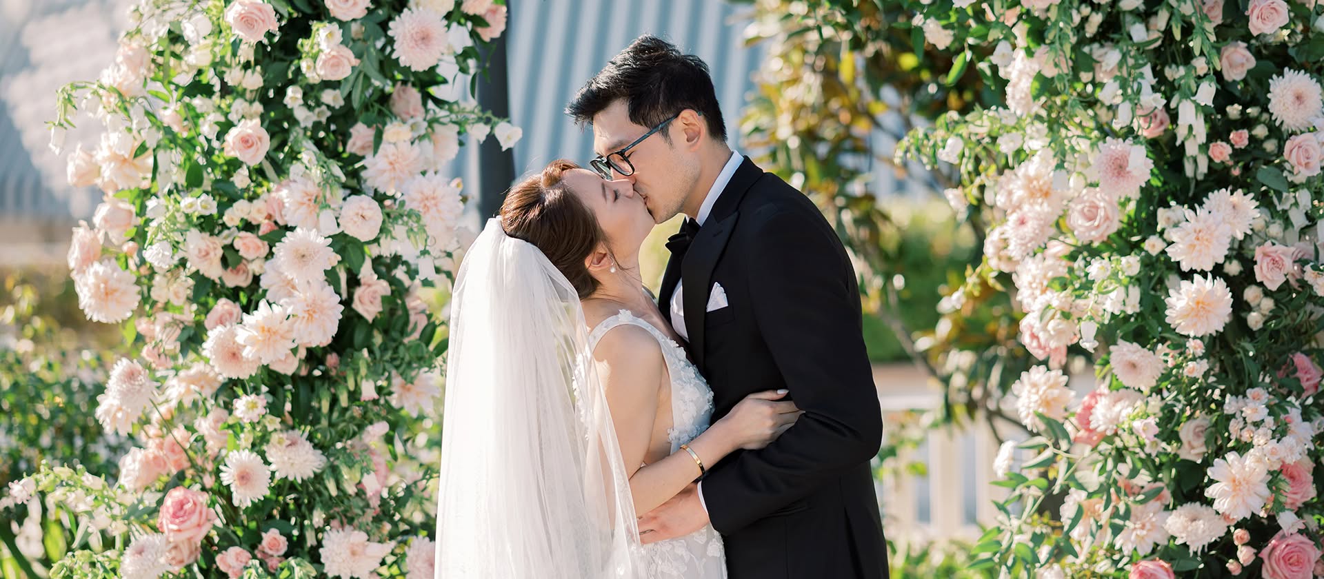 A couple sharing their first kiss beneath a blush floral arch at their San Diego wedding, planned by Bliss Events.