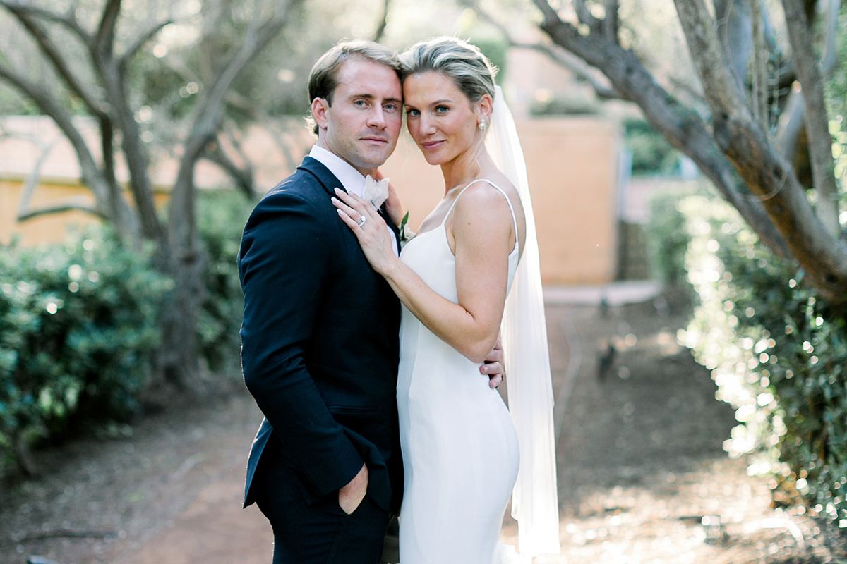 Claire and Collin embracing on a tree-lined garden path, the bride in a ruffled gown and long veil beside the groom in a black tuxedo.