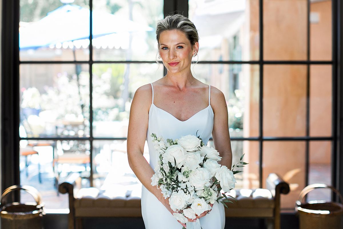 Claire holding a white rose bouquet, standing indoors before tall black-framed windows.