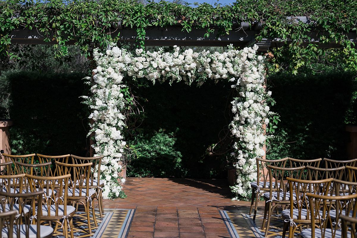 The outdoor ceremony arch covered in white flowers, flanked by gold chairs on a tiled aisle at Rancho Valencia.