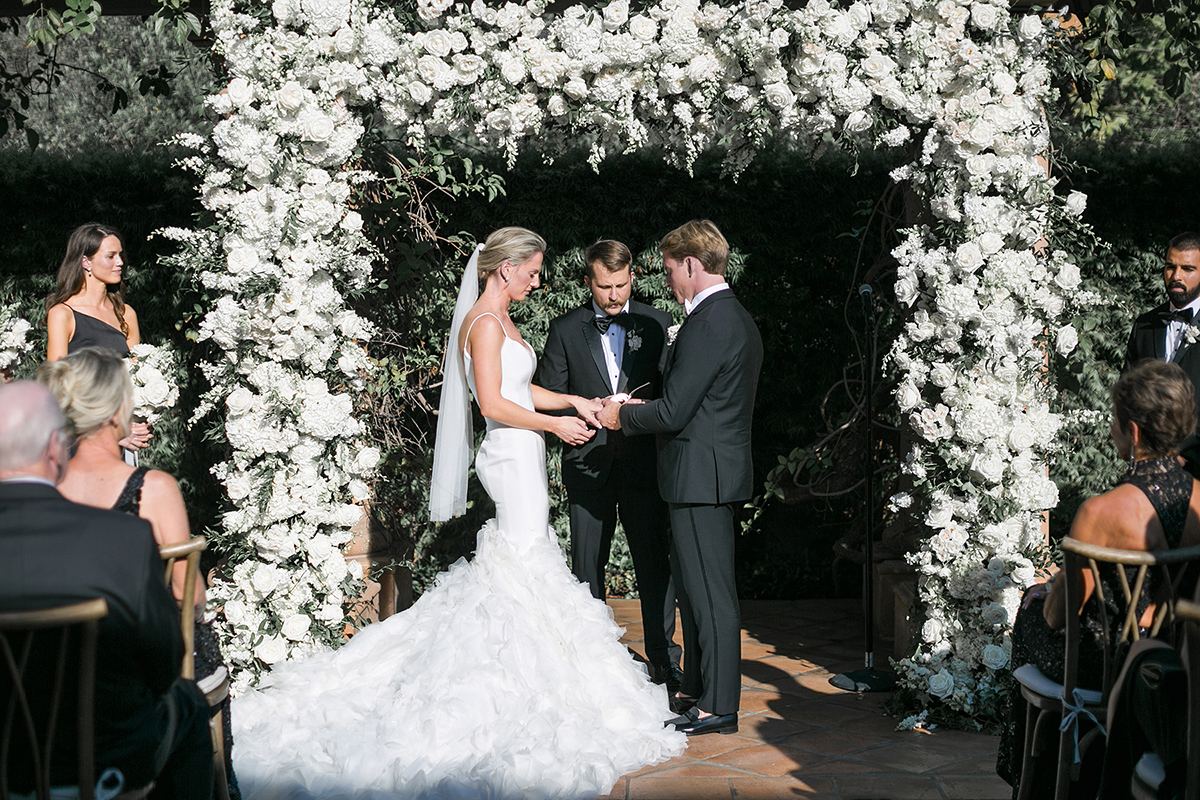Claire and Collin exchanging vows beneath the white floral arch as guests look on.