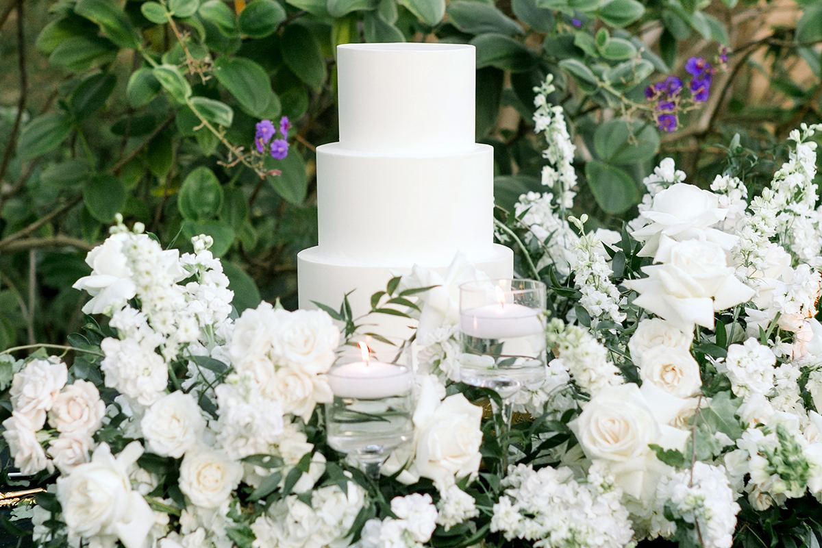 A three-tier white wedding cake on a black-draped table, surrounded by white roses and trailing greenery.