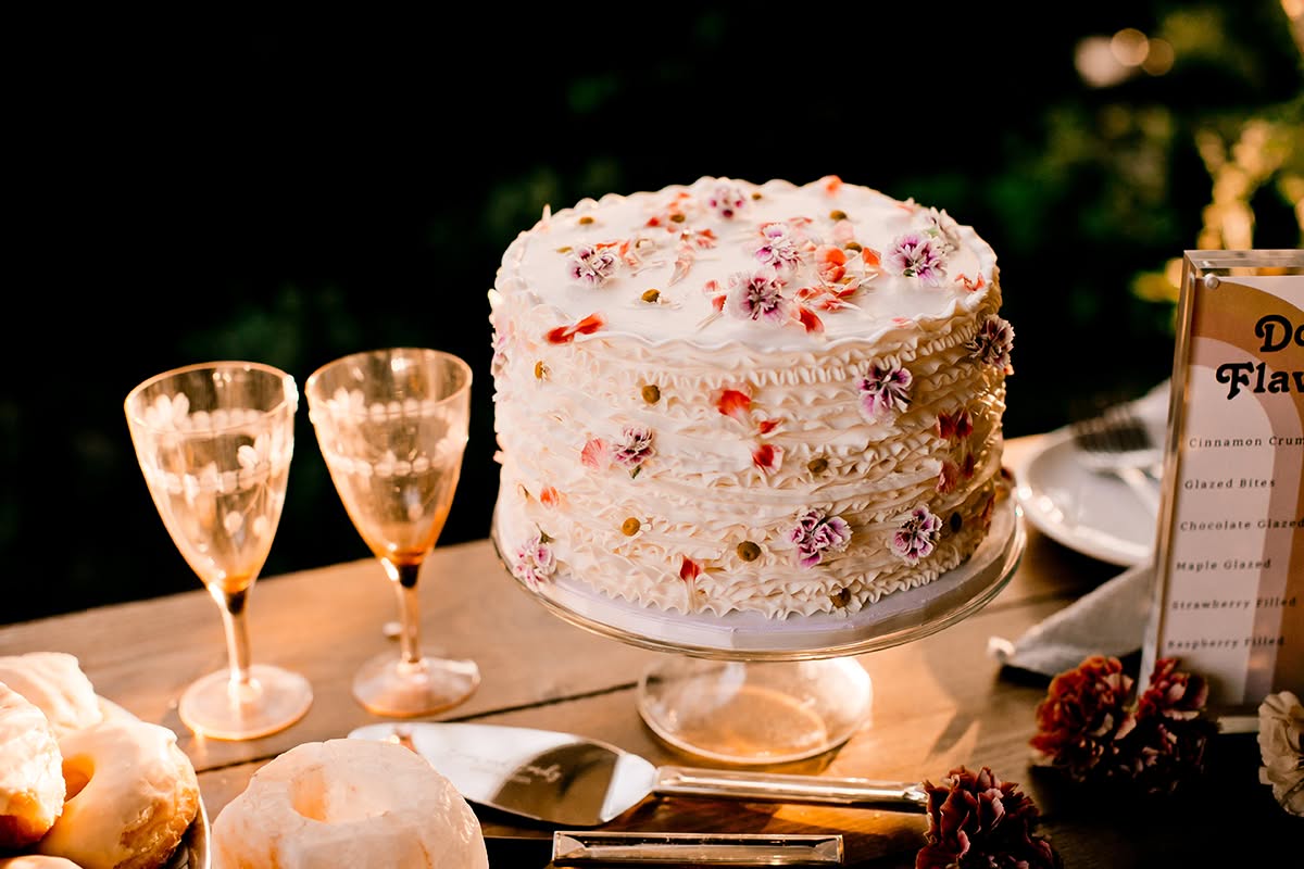 Wedding cake with pressed floral details displayed beside champagne glasses and dessert signage.