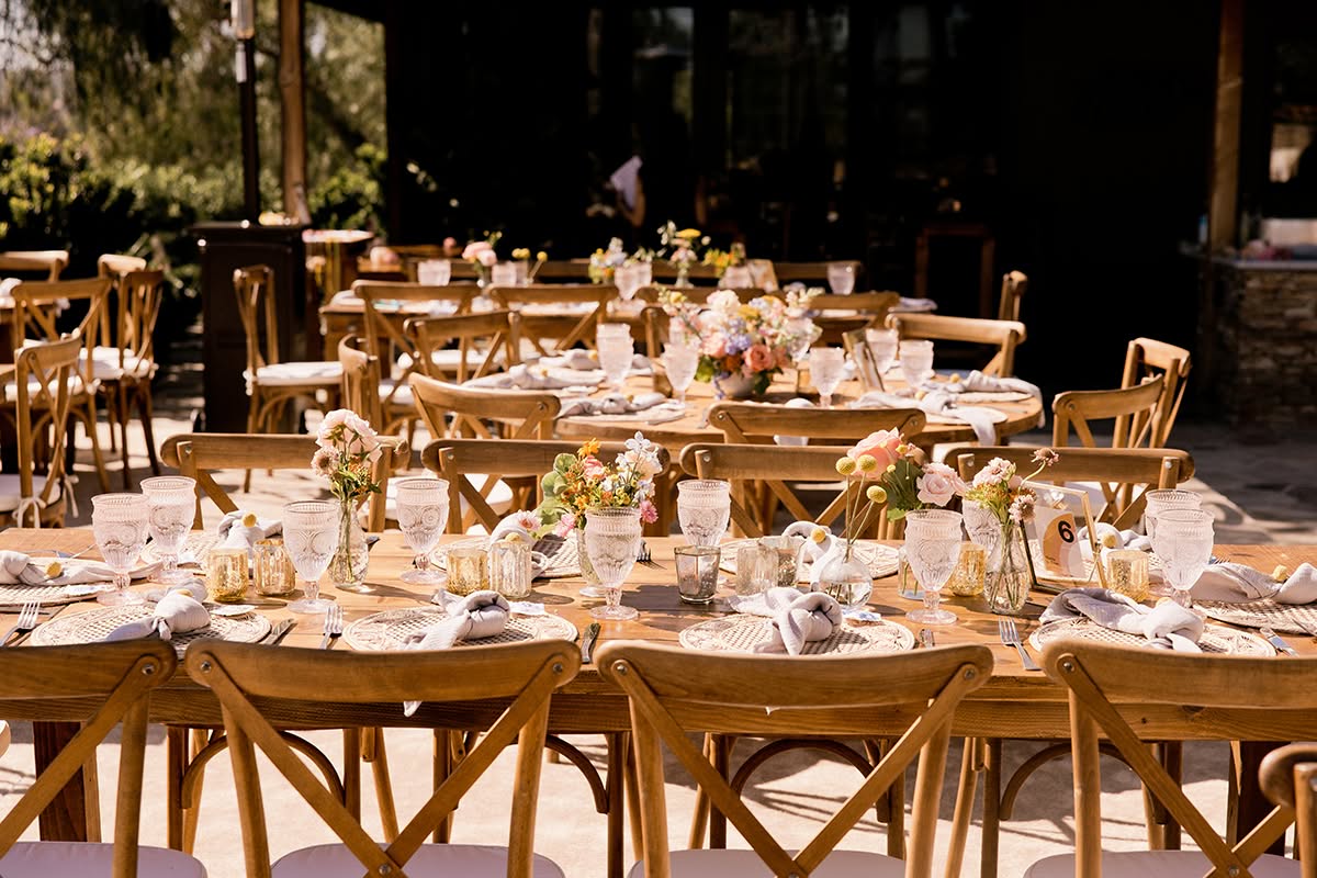 Long outdoor wedding reception table with wood chairs, floral centerpieces, and warm garden light.