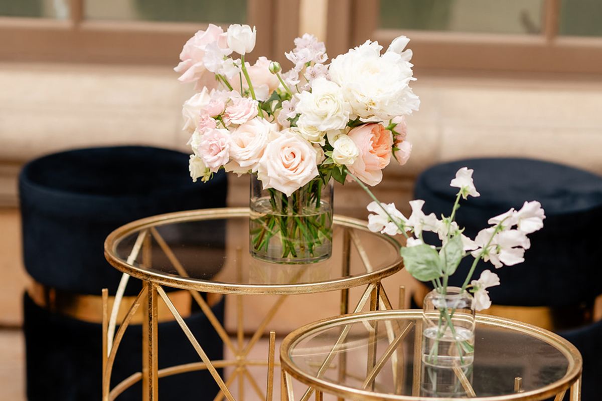 Gold side tables topped with small blush-and-white floral arrangements, beside black velvet seating.
