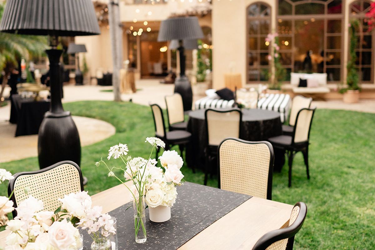 An outdoor dining table with a black runner, white florals, tall black lamps, and palms with the private residence behind.