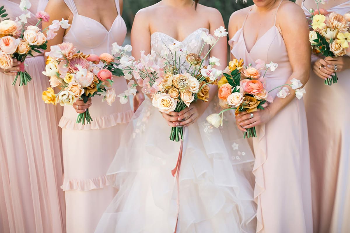 Bride and bridesmaids holding colorful sunset-toned bouquets in soft romantic wedding attire.