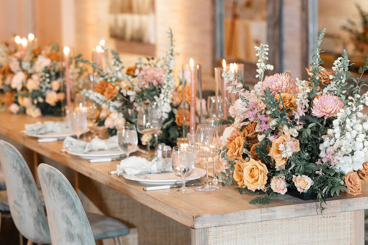 Head table detailed with lush sunset-toned florals, taper candles, and place settings beside gray velvet chairs.