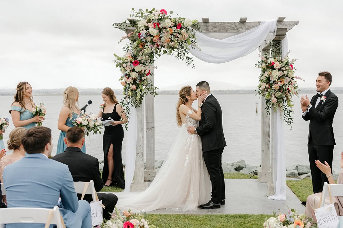 Irma and CJ kiss beneath a floral wedding ceremony arch overlooking the water at Loews Coronado Bay Resort.