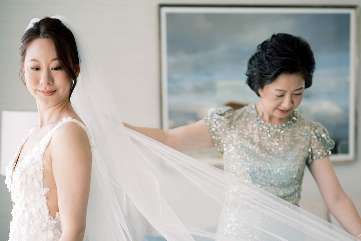 Bride standing indoors as an attendant adjusts her veil before the wedding ceremony.