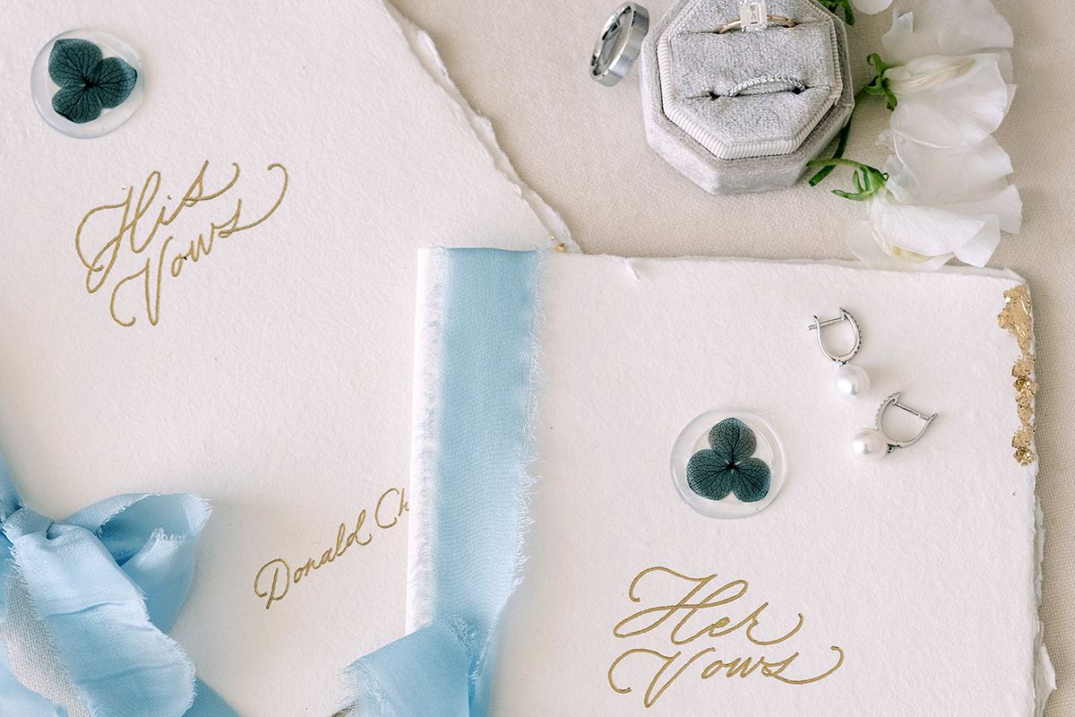 A wedding details flat-lay with His and Her vow books, pale blue silk ribbon, rings, and white florals.