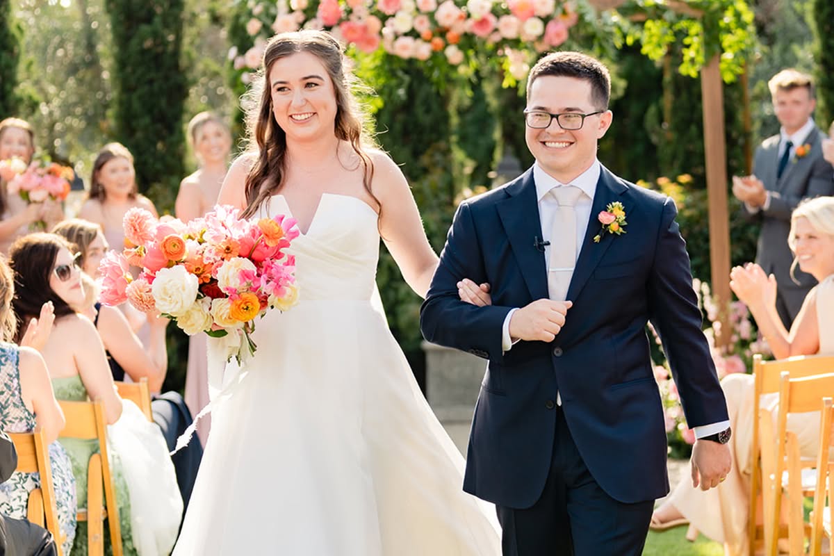 Katie walks down the garden aisle with an escort while holding a bright wedding bouquet.