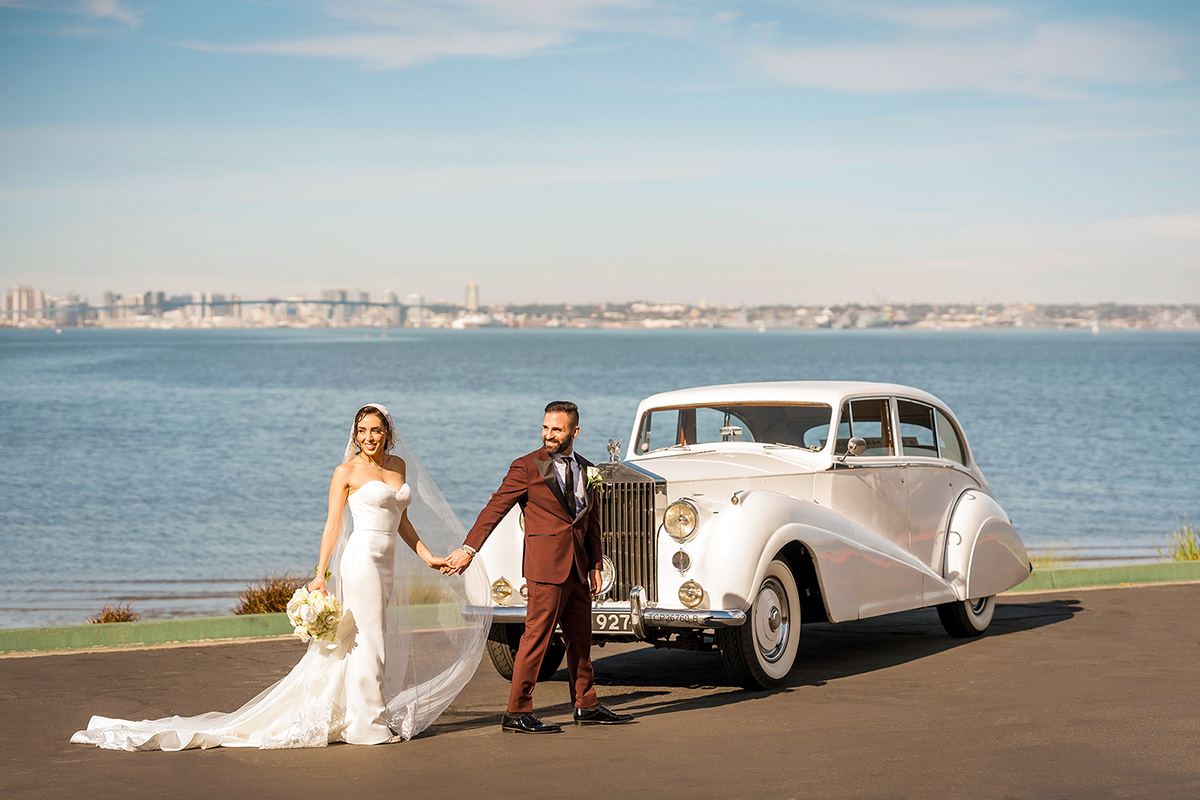 Bride and groom standing beside a vintage white car with Coronado Bay in the background.
