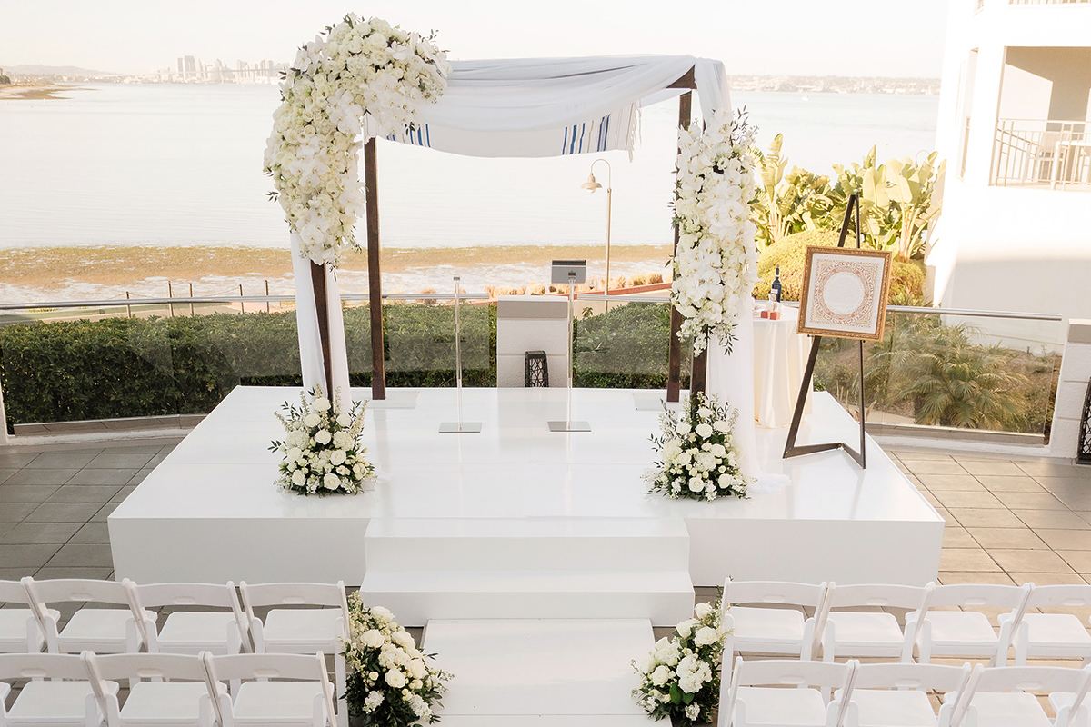 Outdoor ceremony aisle lined with white florals and white chairs, leading to a white floral chuppah overlooking the water.