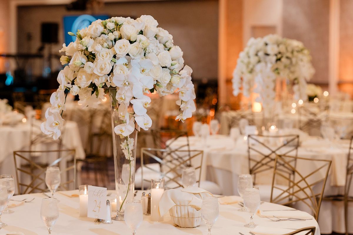 Reception table detail with a tall white floral centerpiece and gold chairs.