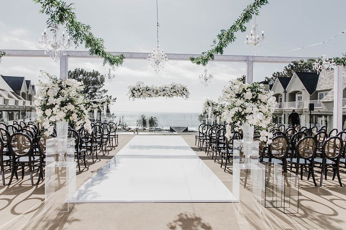 Ocean-view rooftop ceremony aisle with white florals, black chairs, and a crystal chandelier.