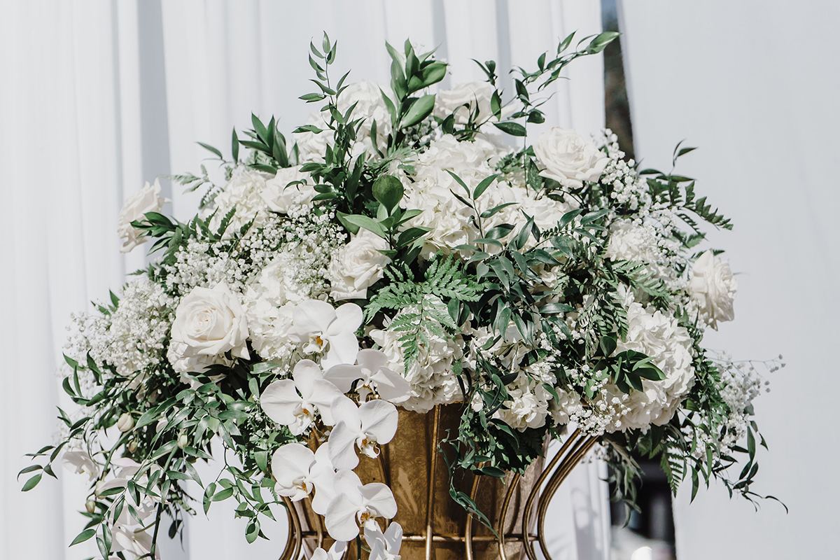 White floral arrangement displayed on a gold stand beside white draping.