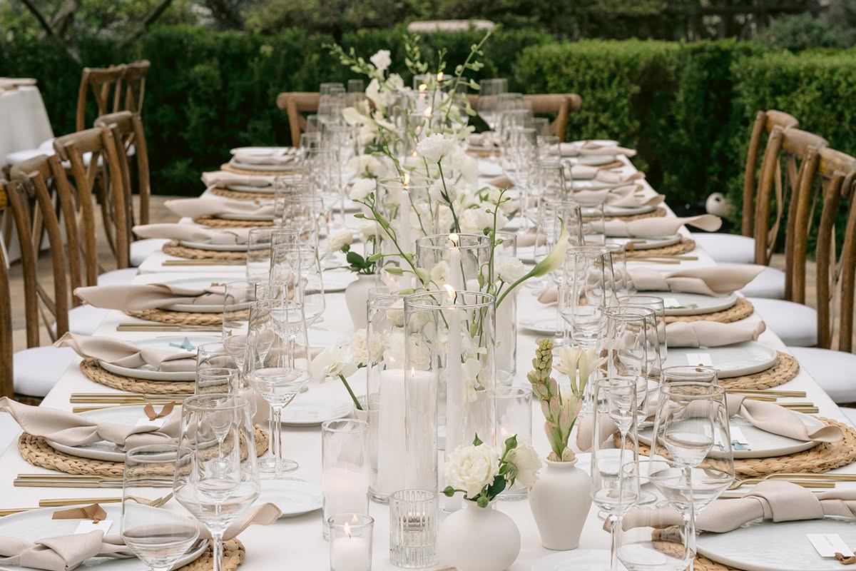 A place setting with a woven charger, linen napkin, and a white floral centerpiece with taper candles.